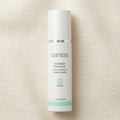 A white cylindrical bottle of Sanitas Antioxidant Moisturizer with a silver band and mint green accents, set against a textured beige linen background.