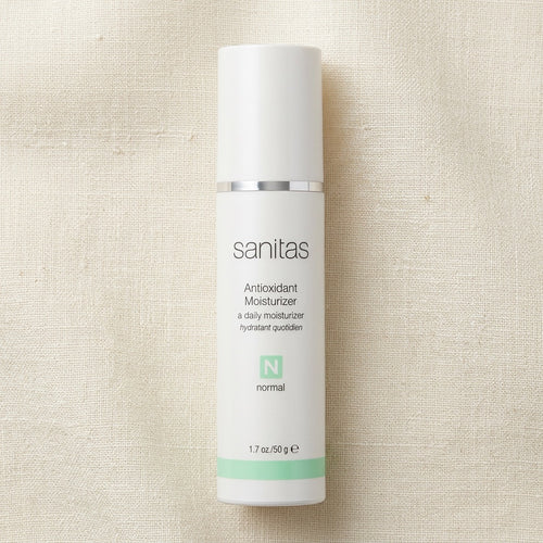 A white cylindrical bottle of Sanitas Antioxidant Moisturizer with a silver band and mint green accents, set against a textured beige linen background.