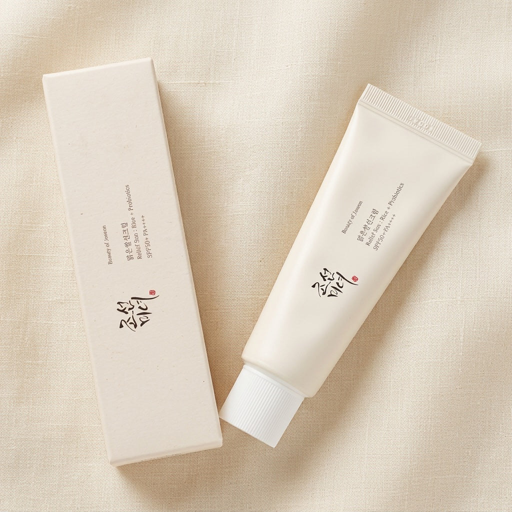 A white cosmetic tube and its matching rectangular box placed on a light beige fabric background. The packaging features black Korean text and a small red seal.