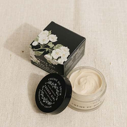 A jar of French Girl body cream with its black lid placed beside it, next to the black product box decorated with white floral illustrations, all arranged on a textured beige fabric background.