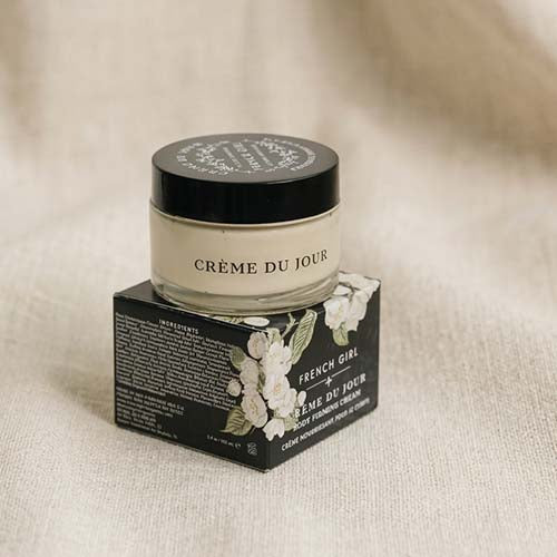 A jar of French Girl x Crème du Jour moisturizing cream sits on top of its black and white floral box against a soft beige linen background.
