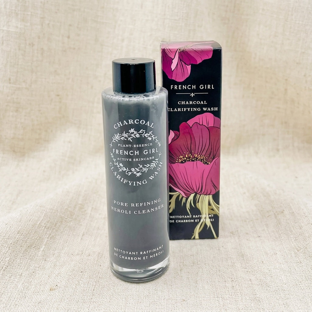 A bottle of French Girl Charcoal Clarifying Wash with a black cap stands next to its retail box on a beige linen background. The bottle contains a gray cleanser, and the box features a black background with large pink flowers.