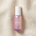A bottle of COOLA Organic Dew Good Illuminating Serum SPF 33, featuring a light purple gradient bottle and a white cap, set against a textured beige linen background.