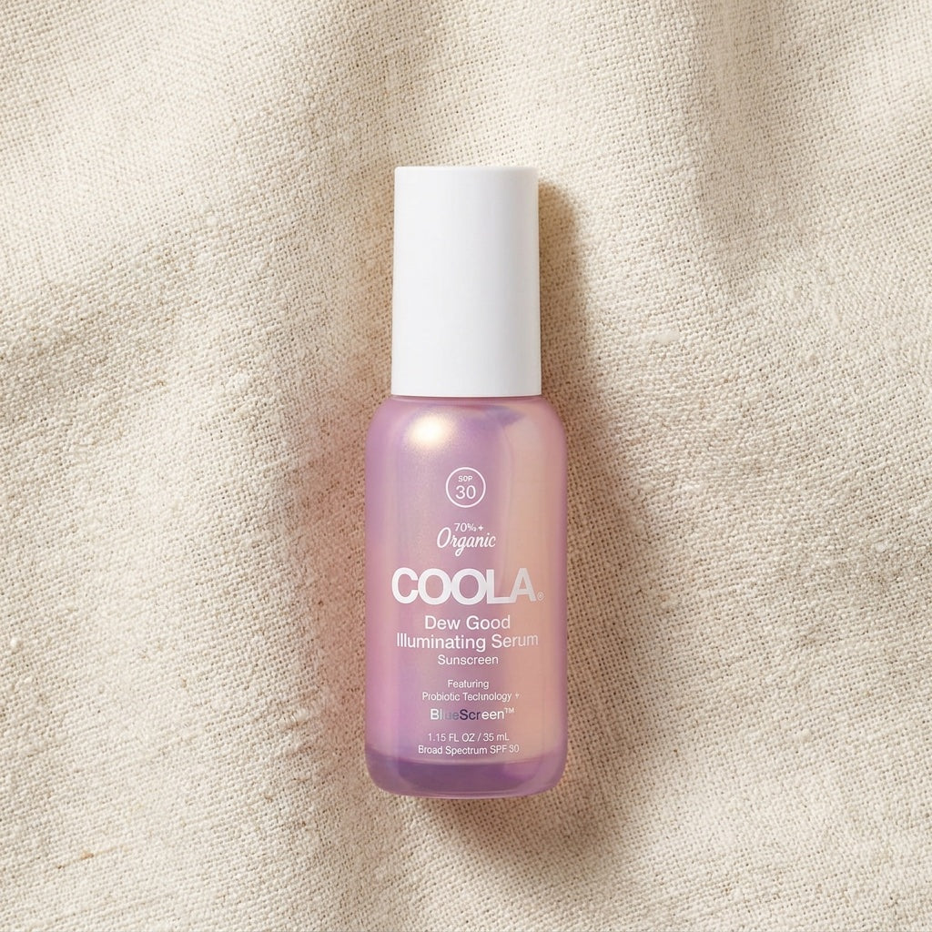 A bottle of COOLA Organic Dew Good Illuminating Serum SPF 33, featuring a light purple gradient bottle and a white cap, set against a textured beige linen background.