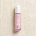 A bottle of COOLA Organic Sun Silk Drops sunscreen, featuring a translucent light pink bottle and a white cap, set against a textured beige linen background.