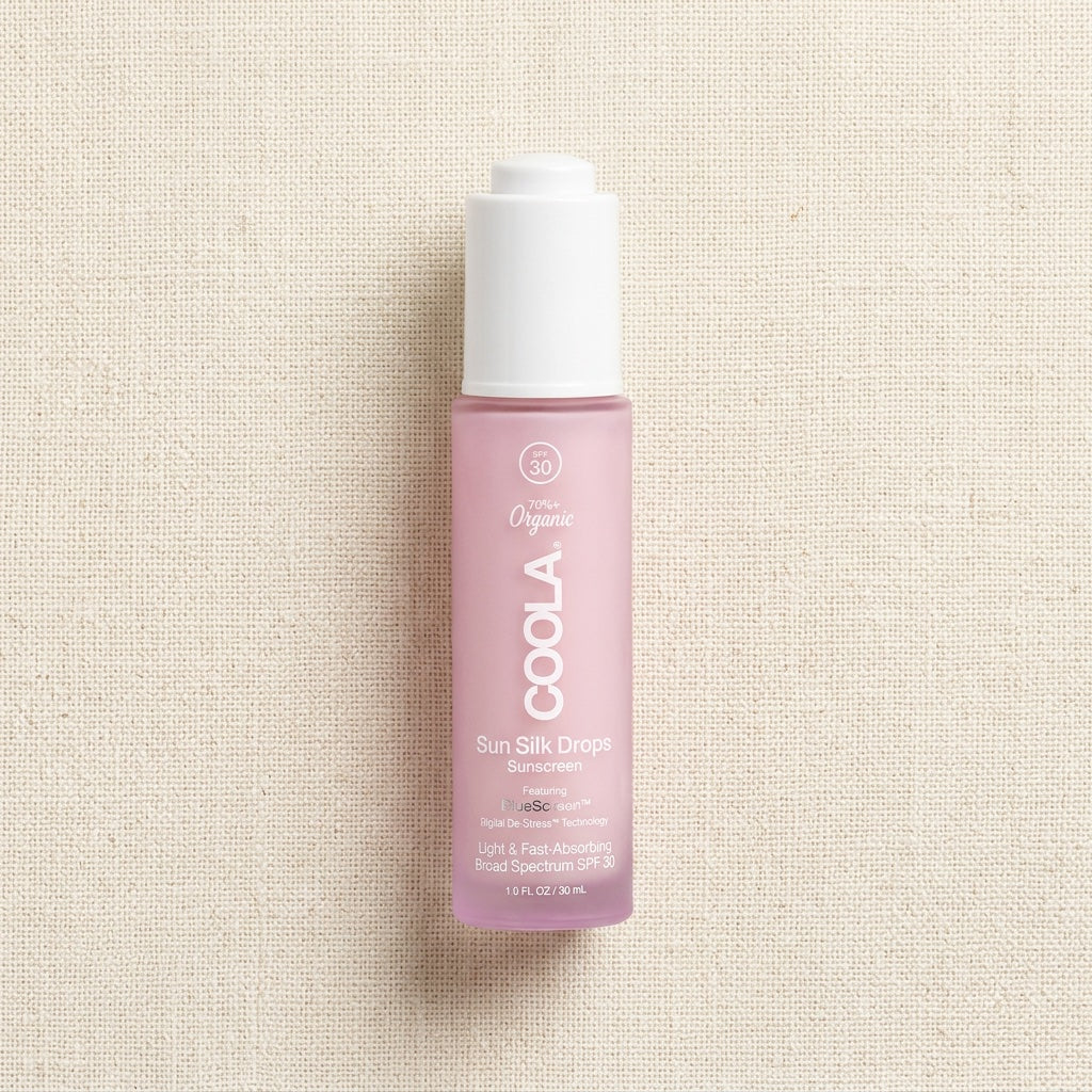 A bottle of COOLA Organic Sun Silk Drops sunscreen, featuring a translucent light pink bottle and a white cap, set against a textured beige linen background.