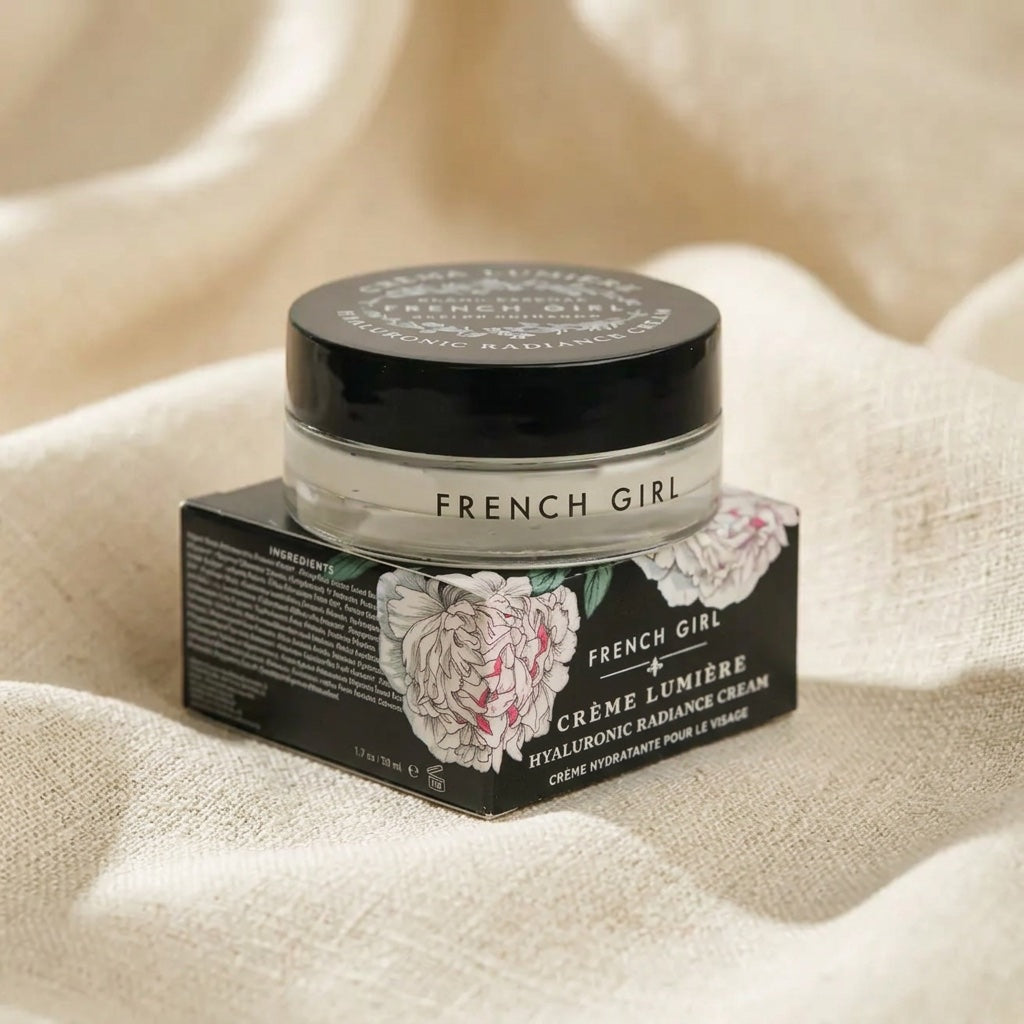 A jar of French Girl Crème Lumère Hyaluconic Radiance Cream sits on top of its black floral product box, set against a soft beige linen fabric background.