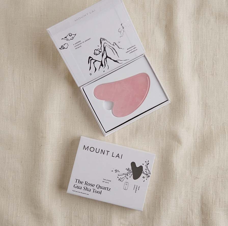 An open white gift box showing a pink rose quartz gua sha tool inside, with a second closed white box placed below it on a beige fabric background. The packaging reads Mount Lai.