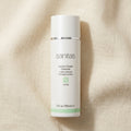 A white bottle of Sanitas Lemon Cream Cleanser with a silver band and a light green accent, set against a textured beige linen background.