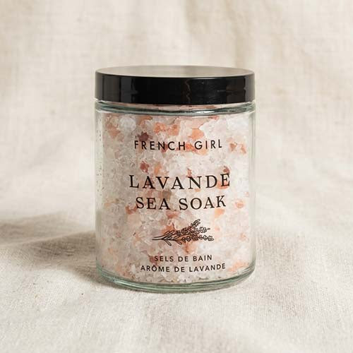 A glass jar of French Girl Lavande Sea Soak bath salts with pink and white crystals, a black lid, and a black printed label, set against a neutral beige linen background.