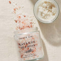 A glass jar labeled 'FRENCH GIRL LAVANDE SEA SOAK' filled with pink and white bath salts, with some spilled onto a beige fabric surface. A small open jar of white scrub or balm sits in the upper right corner.