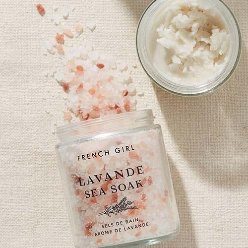 A glass jar labeled 'FRENCH GIRL LAVANDE SEA SOAK' filled with pink and white bath salts, with some spilled onto a beige fabric surface. A small open jar of white scrub or balm sits in the upper right corner.