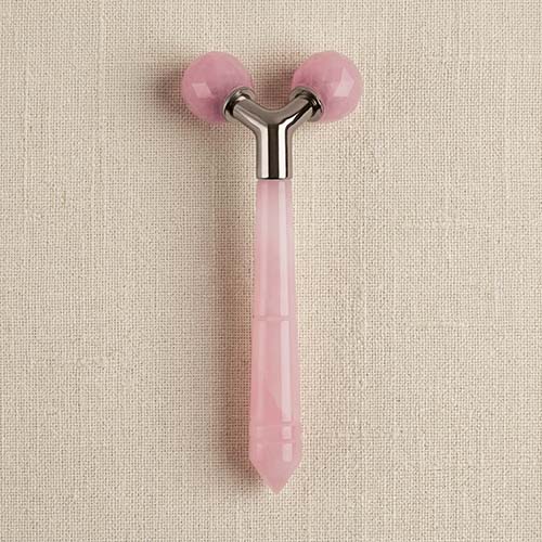 A pink rose quartz facial roller with a silver metal Y-shaped connector and faceted pink gemstone balls, shown on a textured beige linen background.