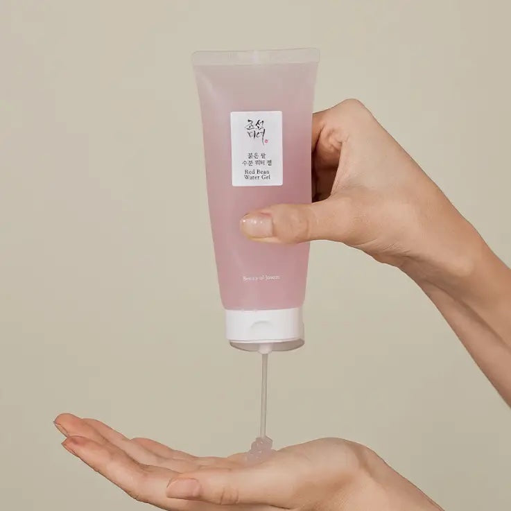 A hand holds a translucent pink cosmetic tube above another hand, with the product being squeezed into the palm. The tube has a white label with Korean text and a white cap.