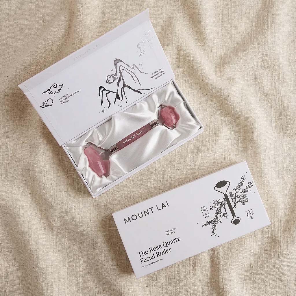 A Mount Lai rose quartz facial roller presented with its packaging on a textured beige linen background. The image shows both the open box containing the pink rose quartz roller and the closed outer box.
