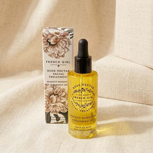 A bottle of French Girl Rose Nectar Facial Treatment cleansing oil with a black dropper cap stands next to its floral-patterned box on a beige fabric surface.