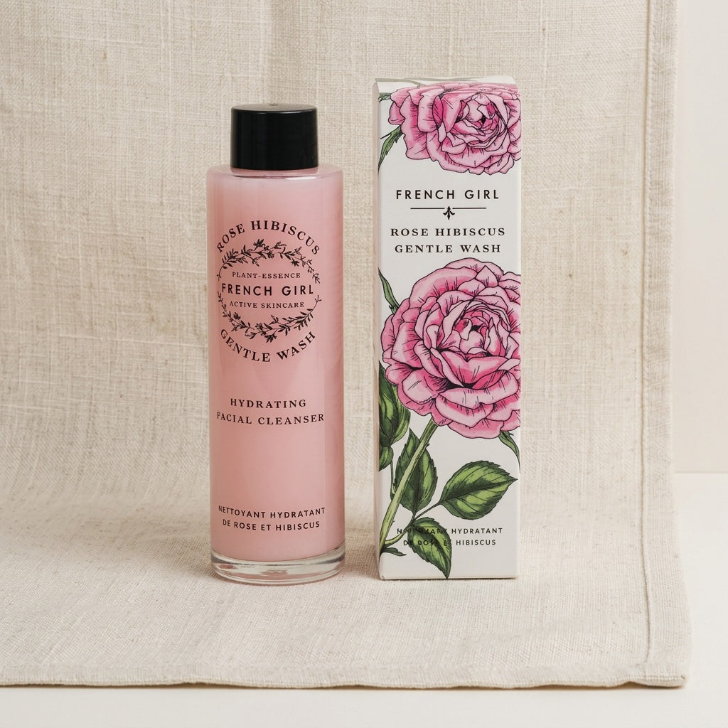 A bottle of French Girl Rose Hibiscus Gentle Wash next to its illustrated packaging box, set against a neutral linen background.