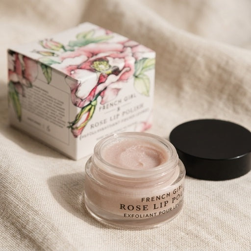 A small clear jar of French Girl Rose Lip Polish with pale pink cream visible inside, shown next to its floral product box and a black lid on a beige fabric surface.