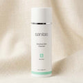 A white cylindrical bottle of Sanitas Sensitive Skin Cleanser with a silver band and mint green accents, set against a draped beige linen background.