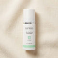 A white bottle of Sanitas Skin Soothing Concentrate with a white cap, featuring a silver band and a light green accent stripe, set against a textured beige linen background.