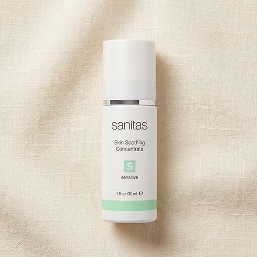 A white bottle of Sanitas Skin Soothing Concentrate with a white cap, featuring a silver band and a light green accent stripe, set against a textured beige linen background.