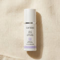 A white bottle of Sanitas Vita C Serum with a silver band and light purple accents, placed on a textured beige linen background.