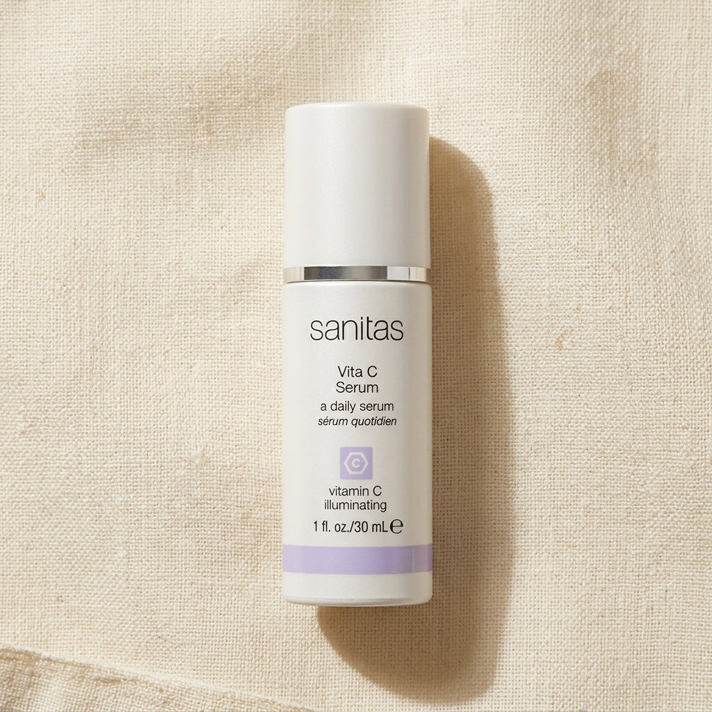 A white bottle of Sanitas Vita C Serum with a silver band and light purple accents, placed on a textured beige linen background.
