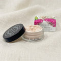 A small clear cosmetic jar labeled 'LAIT DE ROSE' with pale pink powder inside, shown beside its black lid and floral product box on a light beige fabric background.