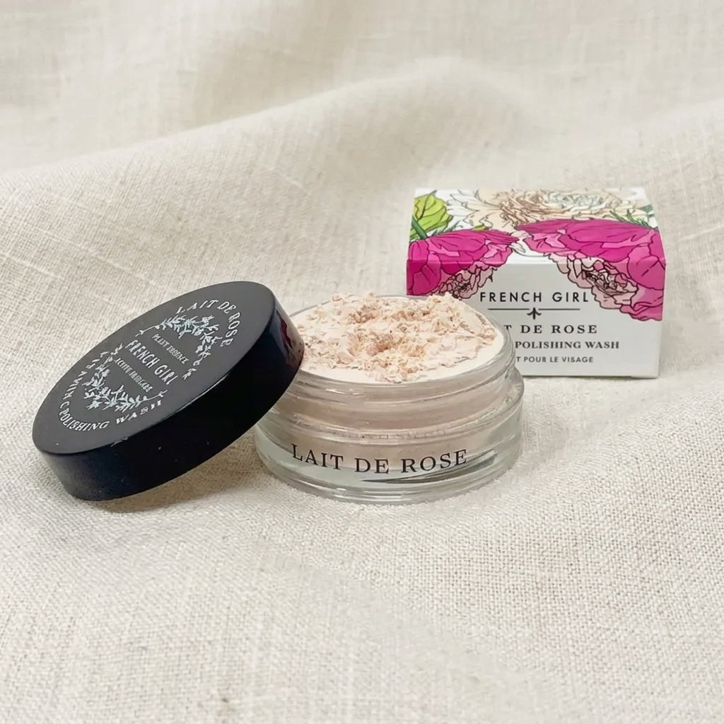 A small clear cosmetic jar labeled 'LAIT DE ROSE' with pale pink powder inside, shown beside its black lid and floral product box on a light beige fabric background.